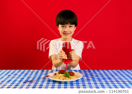 Smiling Korean boy sitting at table and squeezing ketchup sauce on plate with spaghetti and meatballs against red background 114190701