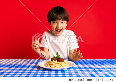 Young boy feeling excited, sitting at table with blue checkered tablecloth and plate of spaghetti and meatballs against red background 114190734