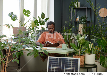 Portrait of adult African American man reading notes in planer while sitting at workplace with lush green plants copy space Portrait of adult African American man reading notes in planer while sitting at workplace with lush green plants copy space 114190866