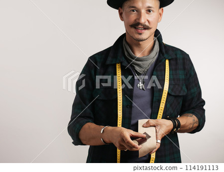 Artisan of leather in creative black hat posing with leather goods in studio. Artisan of leather in creative black hat posing with leather goods in studio. 114191113