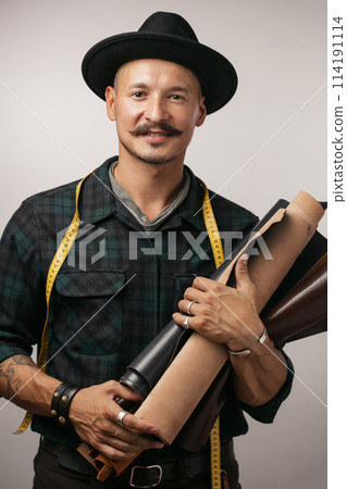 Artisan of leather in creative black hat posing with leather goods in studio. Artisan of leather in creative black hat posing with leather goods in studio. 114191114