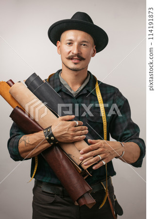 Artisan of leather in creative black hat posing with leather goods in studio. Artisan of leather in creative black hat posing with leather goods in studio. 114191873