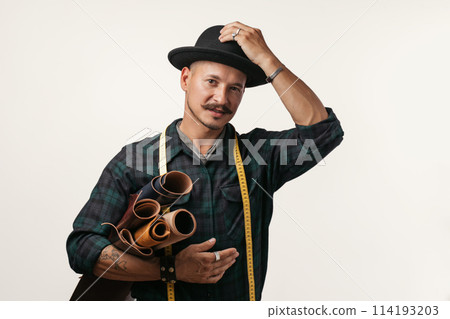Artisan of leather in creative black hat posing with leather goods in studio. Artisan of leather in creative black hat posing with leather goods in studio. 114193203