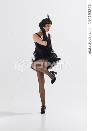 Elegant young woman, dancer styled in Roaring Twenties attire, performing solo Charleston dance isolated over white studio background 114193296