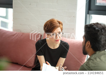 woman is sitting on couch and talking to male indian psychologist while doctor making notes woman is sitting on couch and talking to male indian psychologist while doctor making notes 114194651