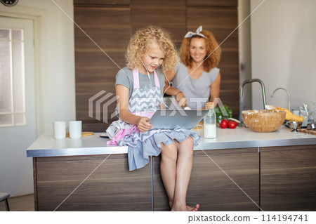 Little girl and her daughter baking cookies at home, reading recipe on laptop. Little girl and her daughter baking cookies at home, reading recipe on laptop. 114194741
