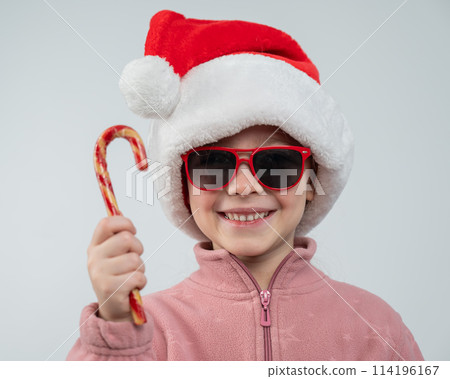 Portrait of a cute Caucasian girl in a Santa Claus hat and sunglasses eating a lollipop on a white background. Portrait of a cute Caucasian girl in a Santa Claus hat and sunglasses eating a lollipop on a white background. 114196167