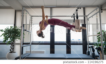 A caucasian woman is engaged in Pilates on a reformer simulator. Hanging position. 114196275