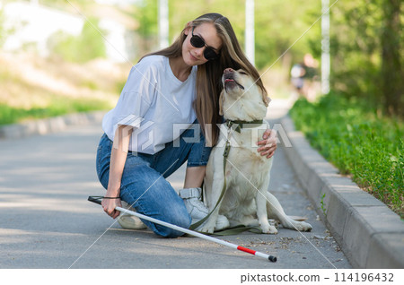 Blind young woman cuddling with guide dog on a walk outdoors. Blind young woman cuddling with guide dog on a walk outdoors. 114196432