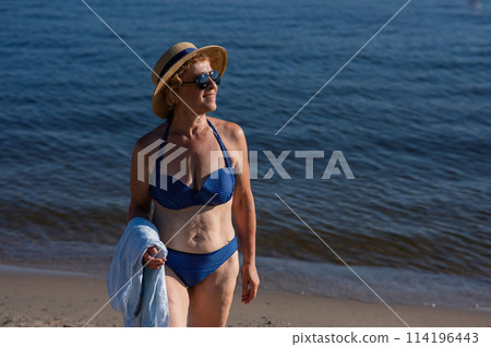 An old woman in a straw hat, sunglasses and a swimsuit is resting on the beach.  114196443