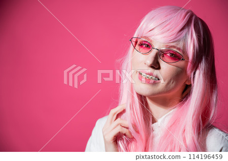 Close-up portrait of a young woman with braces in a pink wig and sunglasses on a pink background. Copy space. Close-up portrait of a young woman with braces in a pink wig and sunglasses on a pink background. Copy space. 114196459