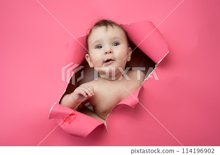 Cute Caucasian newborn baby boy peeks out of a hole in a paper pink background. 114196902