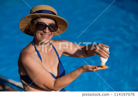 Portrait of an old woman in a straw hat, sunglasses and a swimsuit applying sunscreen to her skin while relaxing by the pool.  114197076