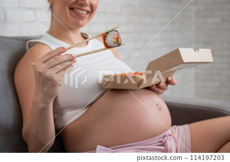 A pregnant woman sits on the sofa and eats rolls from a box. Food delivery.  114197203