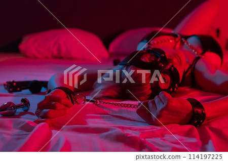 Sexy blonde woman in a leather mask and handcuffs and a collar lies in the bedroom in a red-blue neon light. Role-playing games. Sexy blonde woman in a leather mask and handcuffs and a collar lies in the bedroom in a red-blue neon light. Role-playing games. 114197225