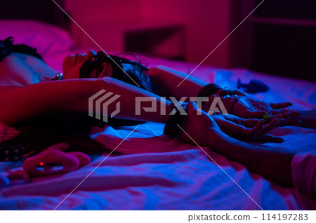 A woman in a leather mask and handcuffs lies on a bed in a blue-red light. Sex toys collection.  114197283