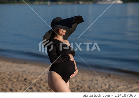 Pregnant woman in a large straw hat and black swimsuit posing on the beach. Pregnant woman in a large straw hat and black swimsuit posing on the beach. 114197360