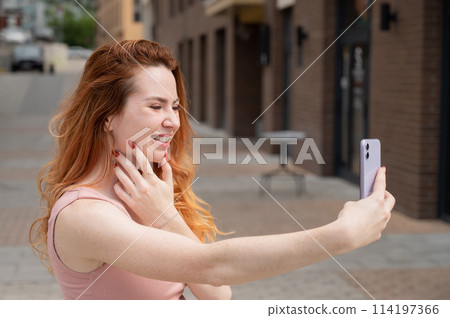 Young woman with braces on her teeth smiles and takes a selfie on a smartphone outdoors. Young woman with braces on her teeth smiles and takes a selfie on a smartphone outdoors. 114197366