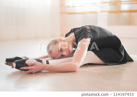 A young woman lies bent over on her feet in a ballet class. Ballerina stretches on the floor. A young woman lies bent over on her feet in a ballet class. Ballerina stretches on the floor. 114197433