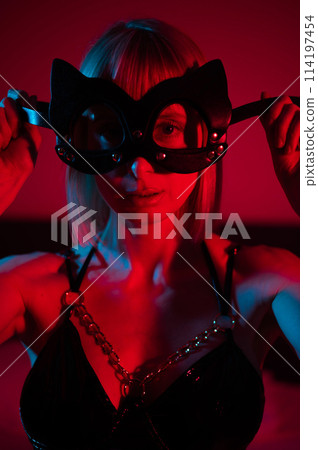 Blonde woman putting on leather mask in bedroom in red blue neon light. Blonde woman putting on leather mask in bedroom in red blue neon light. 114197454