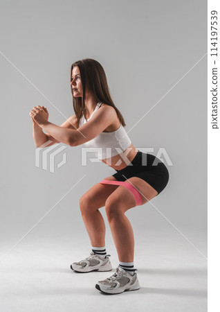 Caucasian woman doing squats with fitness band on white background. Vertical photo. Caucasian woman doing squats with fitness band on white background. Vertical photo. 114197539