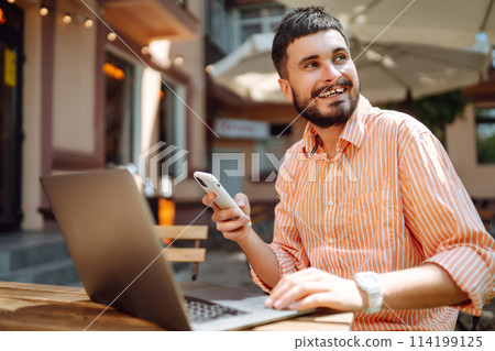 Smiling young man working in cafe on street with laptop and phone. Freelance business concept. 114199125