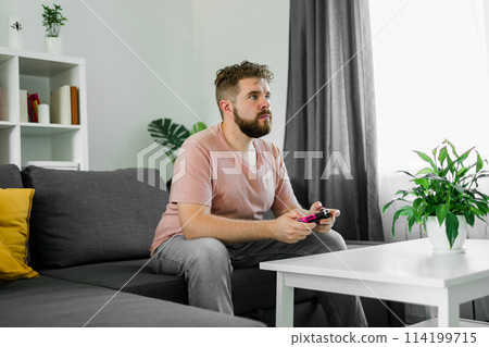 Man playing videogames in living room sitting on sofa. 114199715
