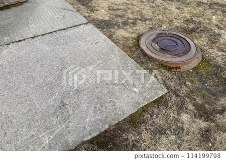 A rusty iron hatch with a pattern is located next to a dry grass pedestrian road during the daytime. Close-up A rusty iron hatch with a pattern is located next to a dry grass pedestrian road during the daytime. Close-up 114199798