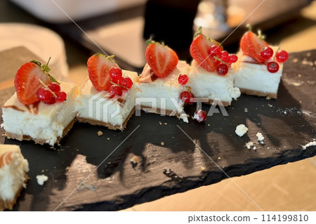 The portions of white cheesecake with strawberry on a black stands on the counter of the restaurant. Close-up The portions of white cheesecake with strawberry on a black stands on the counter of the restaurant. Close-up 114199810