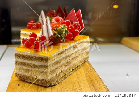 A portion of layered cream yellow cake with red raspberries, red currants on a wooden stand on the counter of the restaurant. Close-up 114199817