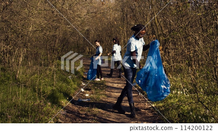 Group of diverse activists picking up the trash and plastic waste, collecting and recycling rubbish in the woods. People doing voluntary work to clean the natural habitat. Camera B. 114200122