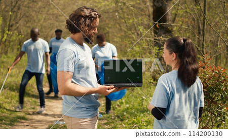 Team of eco friendly activists using pc with isolated display in the woods, volunteering to collect rubbish and plant trees for future generations. Environmental conservation. Camera A. 114200130