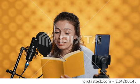 Media star filming vlog at work, reading book to record audiobook, studio background. Voice actor doing content creation while producing digital recording on novel using microphone, camera A 114200143