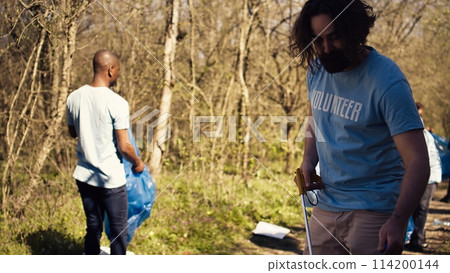 Man activist using tongs to grab garbage and plastic waste, picking up trash and cleaning the forest area. Volunteer sorting rubbish and recycling it, preserving the nature. Camera B. Man activist using tongs to grab garbage and plastic waste, picking up trash and cleaning the forest area. Volunteer sorting rubbish and recycling it, preserving the nature. Camera B. 114200144