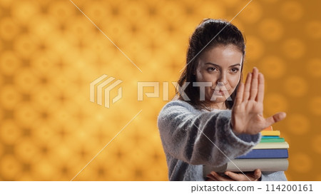 Portrait of stern woman holding stack of books doing stop sign gesturing, studio background. Student with pile of textbooks in arms used for academic learning doing halt hand gesture, camera A 114200161