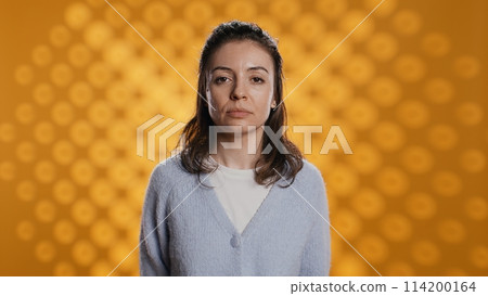 Portrait of relaxed woman posing, looking at camera, isolated over yellow studio background. Caucasian person dressed in casual attire standing, feeling confident, camera B 114200164