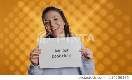 Cheerful book club president holds message urging people to join them, talking about importance of lecture, studio background. Smiling woman inviting bookworms to enlist in her association, camera B Cheerful book club president holds message urging people to join them, talking about importance of lecture, studio background. Smiling woman inviting bookworms to enlist in her association, camera B 114200185