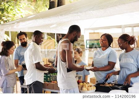 Outdoor food distribution event orchestrated by non-profit, attracting individuals of all races, willing to contribute to welfare of underprivileged. Volunteers supporting the needy with warm meals. Outdoor food distribution event orchestrated by non-profit, attracting individuals of all races, willing to contribute to welfare of underprivileged. Volunteers supporting the needy with warm meals. 114200196