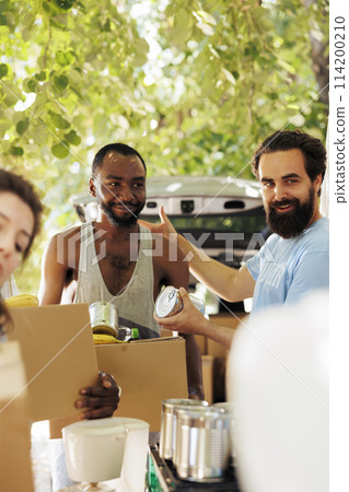 Less fortunate african american man receives canned goods and donation box from caucasian male volunteer. Image showcasing charitable effort aimed to alleviate poverty and hunger within the community. 114200210