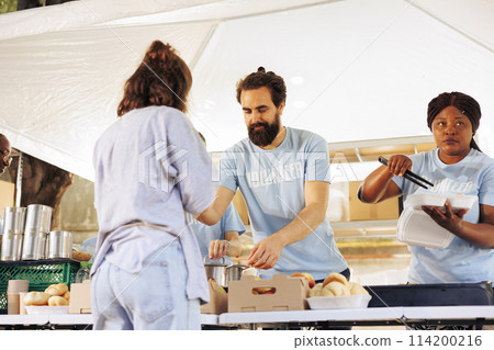 Charitable volunteers distributing free meals to homeless and needy individuals at an outdoor local food bank. Hunger relief team supporting and providing food assistance to the less fortunate. 114200216