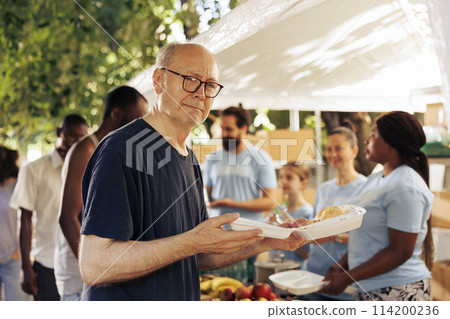 Image showcasing old caucasian homeless man holding his meal from the charitable organization. Group of volunteers at an outdoor food bank helping and feeding the hungry poor and less privileged. Image showcasing old caucasian homeless man holding his meal from the charitable organization. Group of volunteers at an outdoor food bank helping and feeding the hungry poor and less privileged. 114200236