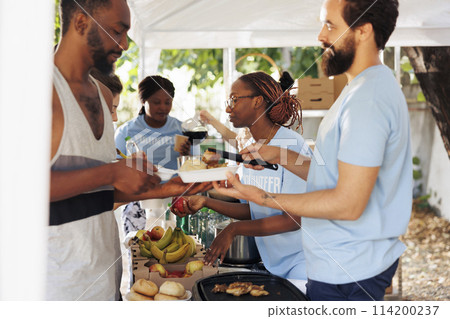 Young African-American and Caucasian voluntary individuals handing out free food and necessities to underprivileged. At homeless shelter, friendly volunteers give hunger relief and humanitarian aid. Young African-American and Caucasian voluntary individuals handing out free food and necessities to underprivileged. At homeless shelter, friendly volunteers give hunger relief and humanitarian aid. 114200237