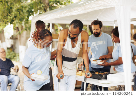Multiethnic charity group handing out free food and nourishments to less fortunate and disabled. Portrait of black woman with blue t-shirt assisting the poor, needy african american man on crutches. 114200241