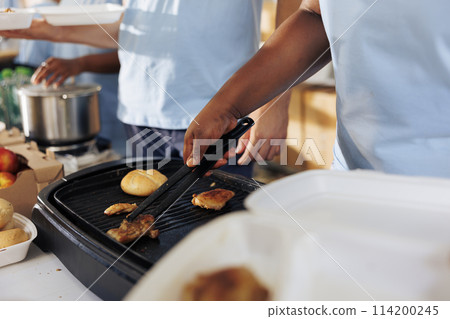 Close-up shot of black woman and caucasian man cooking a free meal for those in need. Image showcasing multiethnic volunteers preparing food for the poor and homeless people at an outdoor food bank. Close-up shot of black woman and caucasian man cooking a free meal for those in need. Image showcasing multiethnic volunteers preparing food for the poor and homeless people at an outdoor food bank. 114200245
