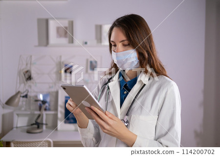 A female doctor in a mask and white uniform uses a tablet in her work. Technologies A female doctor in a mask and white uniform uses a tablet in her work. Technologies 114200702