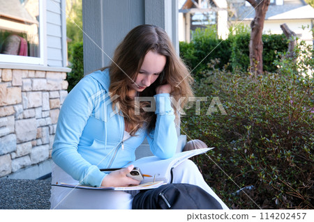 teenager girl does homework on porch of house she writes with pencil in large notebook notebook in folder next to backpack autumn summer sit on threshold beautiful young woman writes draws solves 114202457