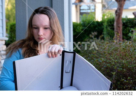 teenage girl with a big white folder in hands crosses out something with pencil writes draws creative person artist young teens outdoors sit by porch have fun holding pen wrong learning doing homework 114202632