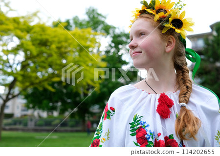 young red-haired girl in a wreath of sunflowers outdoors in park garden in embroidered Ukrainian vyshyvanka shirt with red braid with ribbons in hair Ukraine Peaceful Time of happiness joy and peace 114202655