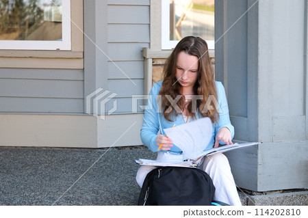 teenager girl does homework on porch of house she writes with pencil in large notebook notebook in folder next to backpack autumn summer sit on threshold beautiful young woman writes draws solves 114202810