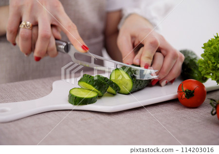 Loving mother teaches daughter to cook vegetable salad, little girl in apron holds knife cutting cucumber on wooden board mom look feel proud share family recipes and experience, pastime hobby concept 114203016